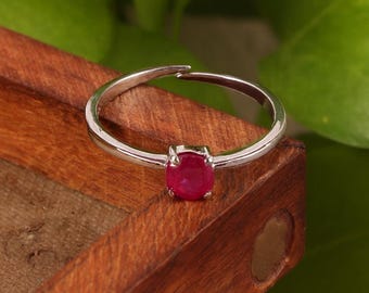 May include: A silver ring with a vibrant red oval gemstone. The ring is set against a wooden surface with green foliage in the background. The gemstone is the focal point, showcasing its color and shape.