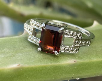 May include: A silver ring with a rectangular, dark red gemstone at the center. The ring features a geometric design with small, clear accent stones on the sides. The ring is set against a green background.