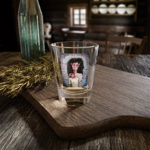 Peut inclure: Un verre à shot en verre transparent orné d'une illustration graphique représentant un personnage pâle aux cheveux noirs avec un visage divisé et une anatomie thoracique exposée. Le texte indique : Bound into this personalized, please fill me up from my despair, what led me to this.