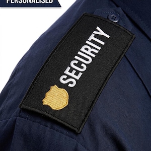May include: A dark blue shirt featuring a rectangular black patch on the shoulder. The patch displays the word SECURITY in white capital letters above a gold-colored shield emblem. A blue banner in the top left corner contains the text PERSONALISED.