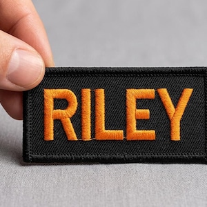 Op de afbeelding: Een rechthoekige zwarte stoffen patch met de naam RILEY geborduurd in opvallende oranje hoofdletters. De patch heeft een gestikte rand en een textuurrijk oppervlak, wat zorgt voor een duidelijk en leesbaar ontwerp voor op textiel.