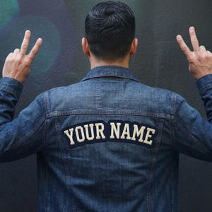 May include: A blue denim jacket featuring a dark navy blue patch on the back with the text YOUR NAME embroidered in white capital letters. The jacket has a classic collar, long sleeves with buttoned cuffs, and visible stitching along the seams.