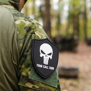 Puede incluir: Un parche negro con forma de escudo fijado en una manga de camuflaje. El parche presenta un gráfico de calavera blanca y el texto "YOUR CALL SIGN" en letras mayúsculas blancas. El diseño está centrado sobre una base de tela rectangular.