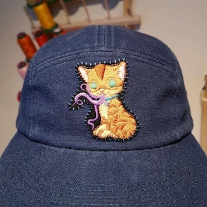 May include: A navy blue baseball cap featuring an embroidered patch of an orange tabby kitten playing with a purple yarn string. The kitten has large green eyes and is outlined with visible blue stitching on the dark fabric of the cap.