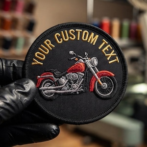 以下が含まれることがあります： 中央に赤いオートバイが刺繍された円形の黒いワッペン。上部の縁に沿って金色の糸でYOUR CUSTOM TEXTという文字が縫い込まれています。ワッペンには黒いステッチの縁取りがあり、細部まで糸でデザインされています。