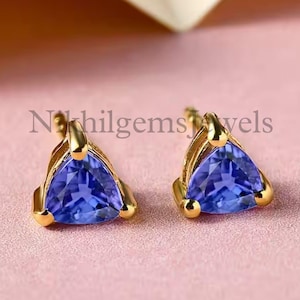 May include: Two gold-tone stud earrings, each with a triangular, deep blue gemstone. The earrings are set on a soft pink surface. The text "Nikhilgemsjewels" is visible.