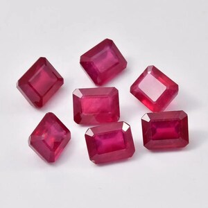 May include: Seven faceted, rectangular-cut rubies. The gemstones are a deep, saturated red color. The rubies are arranged on a white surface, showcasing their clarity and brilliance. Ideal for jewelry making.