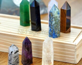 May include: A collection of polished gemstone obelisks in various colors, including green, black, blue, brown, clear, purple, and a black and white speckled stone. The obelisks are displayed on a wooden surface.