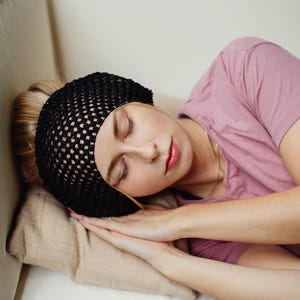 May include: A person resting on a beige pillow, wearing a black net hair cover and a pink shirt. The person is lying on a white couch with their eyes closed, hands clasped near their face.