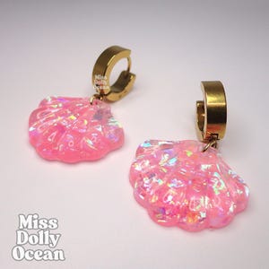Peut inclure: Une paire de boucles d'oreilles créoles avec des pendentifs en forme de coquillage rose à la finition irisée et scintillante. Les anneaux sont en métal doré. Le texte Miss Dolly Ocean est imprimé sur les breloques et dans le coin.