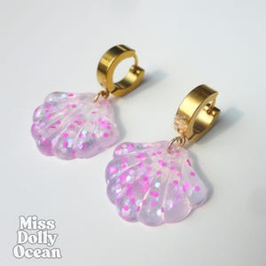 Peut inclure: Une paire de boucles d'oreilles créoles avec des breloques en forme de coquillage en résine violette translucide incrustée de paillettes roses. Les anneaux sont en métal doré. Le texte Miss Dolly Ocean est visible sur les breloques.