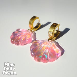 Peut inclure: Boucles d'oreilles coquillage roses iridescentes avec des créoles dorées. Les boucles d'oreilles ont un effet nacré, captant la lumière. Le texte "Miss Dolly Ocean" est visible en bas à gauche.