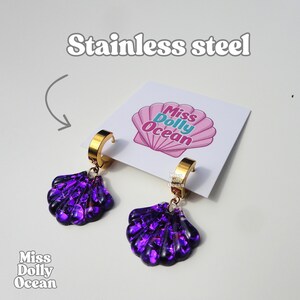 Peut inclure: Une paire de boucles d'oreilles créoles en acier inoxydable doré avec des breloques en forme de coquillage violet à la finition scintillante. Les boucles sont fixées sur une carte blanche avec le texte Miss Dolly Ocean et un motif coquillage.