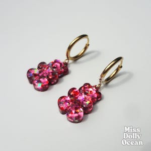 Peut inclure: Une paire de boucles d'oreilles créoles avec des breloques en forme d'oursons en gélatine rose translucide remplies de paillettes irisées. Les breloques sont suspendues à des anneaux dorés. Le texte Miss Dolly Ocean est visible.