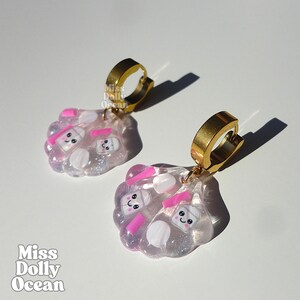 Peut inclure: Une paire de boucles d'oreilles créoles dorées avec des breloques translucides en forme de coquillage. Les breloques contiennent des accents circulaires blancs et roses et de petites figurines de tasses souriantes. Le texte Miss Dolly Ocean est visible.