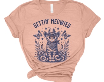 May include: A peach-colored t-shirt featuring a graphic of a cat wearing a cowboy hat and bandana, surrounded by butterflies, stars, and floral elements. The text "GETTIN' MEOWIED" is printed above the cat. A casual, short-sleeved tee.