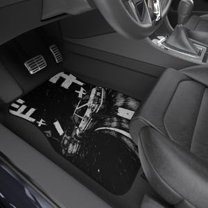 May include: A black car floor mat featuring a high-contrast, black and white graphic illustration of a car drifting on a road. The design includes stylized Japanese characters and motion blur effects, creating a dynamic scene on the rectangular mat.