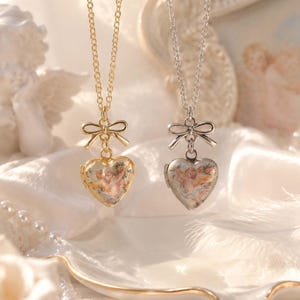 Puede incluir: Dos relicarios en forma de corazón que cuelgan de cadenas delicadas, uno en oro y otro en plata. Cada relicario presenta un detalle de lazo en la parte superior y una ilustración impresa de un querubín alado rodeado de flores en el frente.