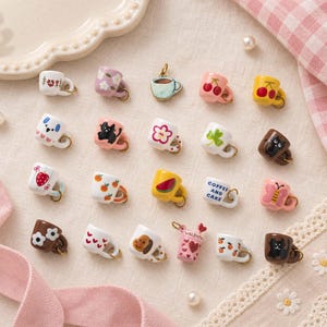 May include: A collection of twenty miniature ceramic mug charms, each featuring unique hand-painted designs including cats, flowers, fruits, hearts, and text. Each tiny mug is equipped with a small gold-toned metal jump ring for attachment.