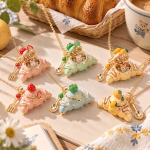 Peut inclure: Une collection de six colliers dorés avec des pendentifs en forme de croissant qui s'ouvrent pour révéler de petites figurines d'animaux. Chaque pendentif est orné de fruits colorés et accompagné d'une mini cuillère ou fourchette assortie.