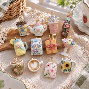 Könnte beinhalten: Eine Sammlung von zwölf Miniatur-Keramikanhängern mit verschiedenen Motiven, darunter eine geblümte Kaffeetasse, eine Teekanne mit Herz, ein Getränk mit Krone, eine Sodaflasche, ein Zitronenbecher, ein Milchkarton, ein Geschenk und verschiedene Tassen.