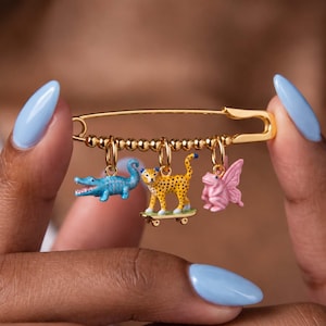 May include: A gold-toned safety pin brooch featuring a row of small metallic beads and three dangling charms: a light blue alligator, a yellow and black spotted leopard standing on a miniature skateboard, and a pink frog with wings.