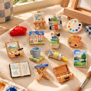 Puede incluir: Una colección de dijes en miniatura con temática artística que incluye una boina roja, un caballete con una pintura, tubos de pintura, una paleta, un estudio de arte, una taza con pinceles, un cuaderno, un taburete y cajas de pintura abiertas.