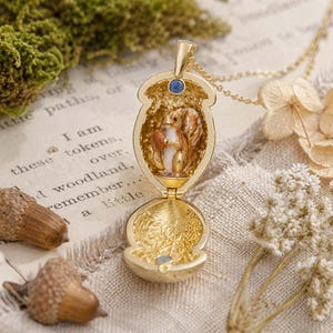 Puede incluir: Un colgante de medallón de metal dorado con forma de bellota, con un diseño articulado que se abre para revelar una figura de ardilla en miniatura sosteniendo una nuez. El interior del medallón muestra un patrón de hojas texturizado y una piedra azul.