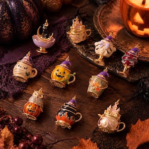 May include: A collection of ten metal charms featuring Halloween-themed designs including a black cat on a crescent moon, a haunted castle teapot, a ghost, a mummy mug, a jack-o'-lantern, an owl on a pumpkin, and a cauldron with orange flames.