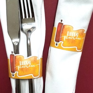 Könnte beinhalten: Zwei weiße Servietten mit orangefarbenen und gelben 'Happy Purim'-Rollenmustern. Die Servietten sind um ein silbernes Messer und eine Gabel gewickelt.