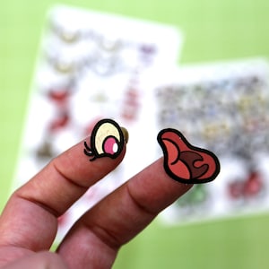 May include: Two colorful stickers, one a pink and white eye with eyelashes and the other a red and black tongue, are shown on two fingers.