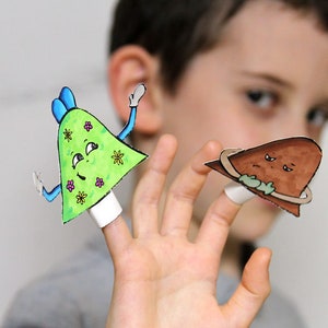 May include: A child's hand wearing two finger puppets. One puppet is green with a flower pattern and a smiling face. The other puppet is brown with a sad face and a ring around it.