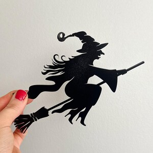 May include: A black silhouette of a witch on a broomstick. The witch wears a pointed hat and flowing robes. The broomstick has detailed bristles. A hand with red nail varnish holds the witch cutout against a white background.