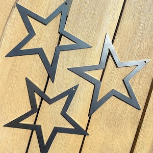 May include: Three dark grey metal star cutouts with a hole at the top for hanging. The stars are arranged on a light brown wooden surface. The star shapes have five points and are open in the centre.
