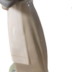 K&ouml;nnte beinhalten: Porzellanfigur, die eine Person in einem langen Mantel darstellt. Die Figur ist haupts&auml;chlich in Beige-, Grau- und Wei&szlig;t&ouml;nen gehalten, mit einem hellgr&uuml;nen Sockel. Die Figur hat eine gl&auml;nzende Oberfl&auml;che und scheint ein Dekorationsartikel zu sein.