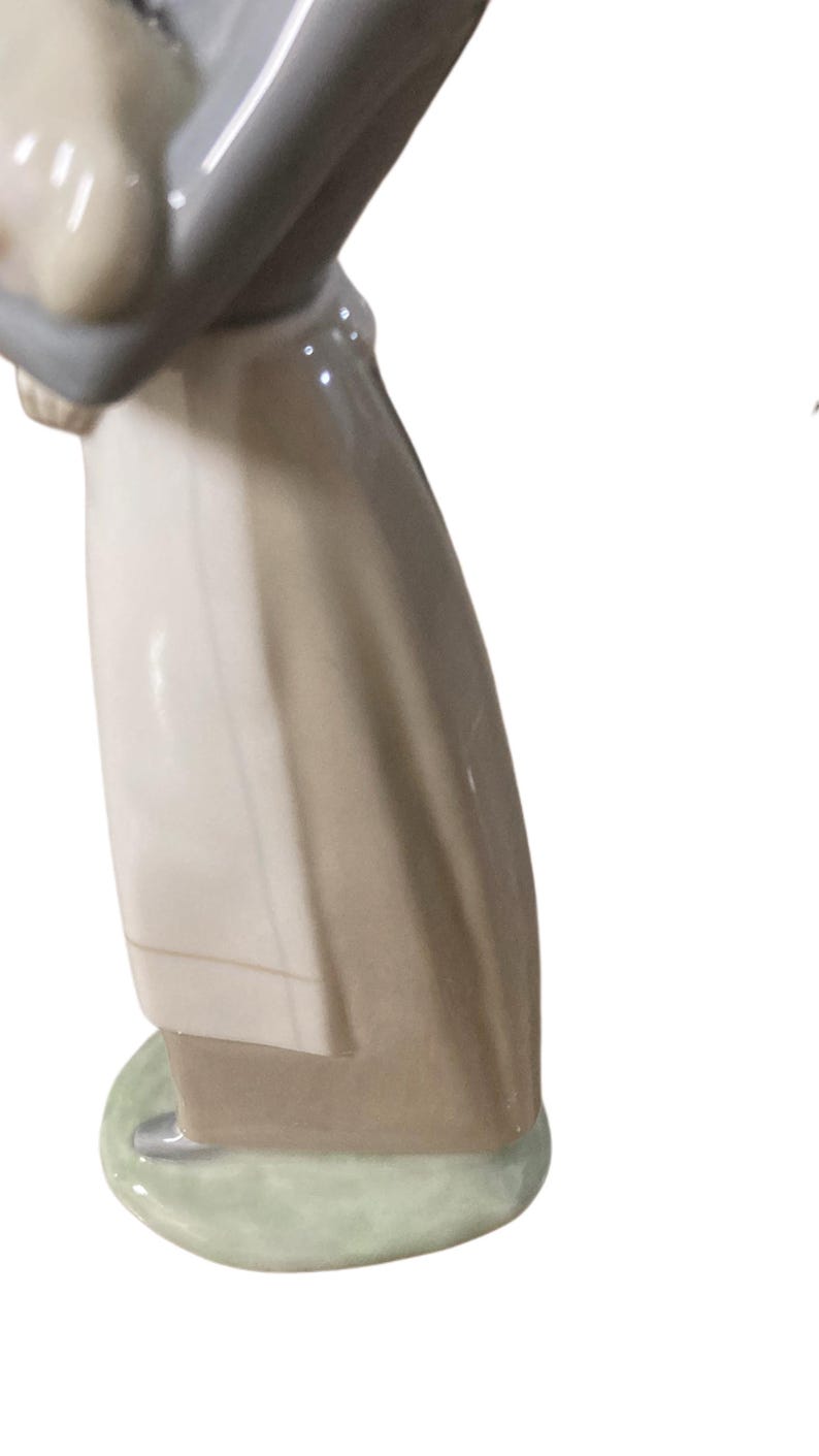 K&ouml;nnte beinhalten: Porzellanfigur, die eine Person in einem langen Mantel darstellt. Die Figur ist haupts&auml;chlich in Beige-, Grau- und Wei&szlig;t&ouml;nen gehalten, mit einem hellgr&uuml;nen Sockel. Die Figur hat eine gl&auml;nzende Oberfl&auml;che und scheint ein Dekorationsartikel zu sein.
