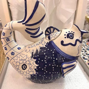 May include: A ceramic fish-shaped pitcher featuring a glossy white and deep blue color scheme. The body is decorated with intricate hand-painted patterns including small flowers, a grid design, and white dots on a dark blue background. The handle is formed by the curved tail, and the spout is integrated into the fish head design.