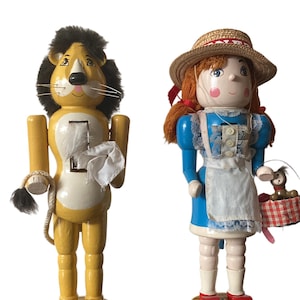 May include: Two wooden nutcrackers from The Wizard of Oz. One is a yellow lion with a black mane and a white cloth. The other is a girl in a blue dress, straw hat, and a basket with a dog.