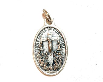 May include: Silver-toned oval pendant with the text "HOLY SOULS IN PURGATORY". The pendant features a crucifix with figures below and on either side. A small loop is at the top for attaching to a chain.