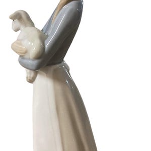 K&ouml;nnte beinhalten: Porzellanfigur einer jungen Person, die ein Lamm h&auml;lt. Die Figur tr&auml;gt ein langes, flie&szlig;endes Kleid mit einem hellblauen Oberteil. Das Lamm ist wei&szlig; und die Figur hat eine gl&auml;nzende Oberfl&auml;che.