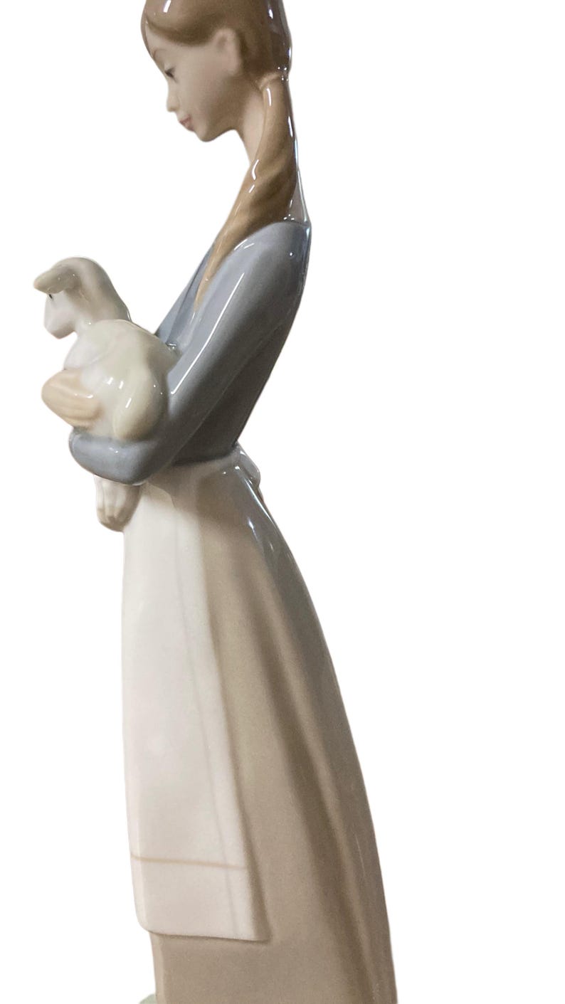 K&ouml;nnte beinhalten: Porzellanfigur einer jungen Person, die ein Lamm h&auml;lt. Die Figur tr&auml;gt ein langes, flie&szlig;endes Kleid mit einem hellblauen Oberteil. Das Lamm ist wei&szlig; und die Figur hat eine gl&auml;nzende Oberfl&auml;che.