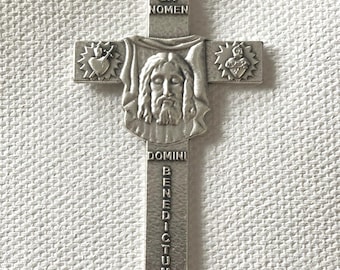 Może przedstawiać: Srebrny wisiorek z krzyżem religijnym. Krzyż przedstawia wizerunek Jezusa Chrystusa z napisem "SIT NOMEN DOMINI BENEDICTUM". Krzyż ma pętlę u góry do zawieszenia.