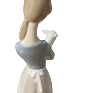 K&ouml;nnte beinhalten: Porzellanfigur, die eine Frau mit einem Lamm zeigt. Die Frau tr&auml;gt ein hellblaues Oberteil und ein langes, cremefarbenes Kleid. Ihr Haar ist zur&uuml;ckgebunden. Das Lamm ist wei&szlig;. Die Figur ist ein Dekorationsartikel.