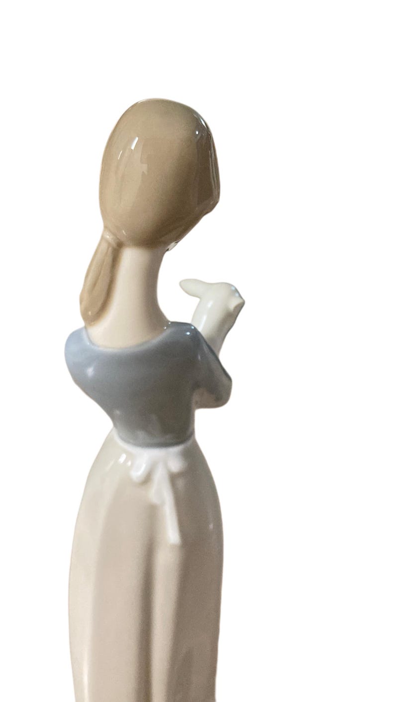 K&ouml;nnte beinhalten: Porzellanfigur, die eine Frau mit einem Lamm zeigt. Die Frau tr&auml;gt ein hellblaues Oberteil und ein langes, cremefarbenes Kleid. Ihr Haar ist zur&uuml;ckgebunden. Das Lamm ist wei&szlig;. Die Figur ist ein Dekorationsartikel.