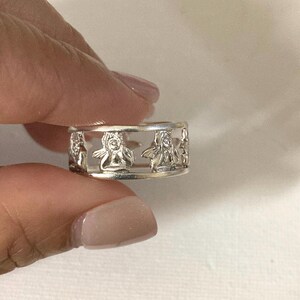 May include: A silver-toned ring featuring a cut-out design that displays a repeating pattern of small, winged figures. The band is constructed with two thin, parallel horizontal rails that connect the series of figures around the entire circumference.