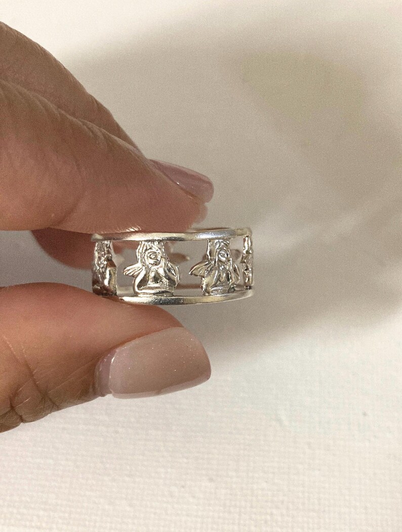 May include: A silver-toned ring featuring a cut-out design that displays a repeating pattern of small, winged figures. The band is constructed with two thin, parallel horizontal rails that connect the series of figures around the entire circumference.