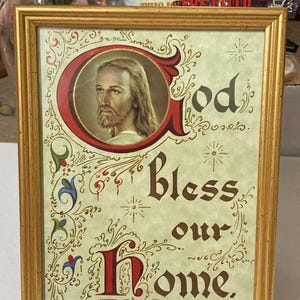 May include: A framed print with a gold-colored frame. The print features an image of Jesus Christ and the words "God Bless our Home" in ornate calligraphy. Decorative floral and star-like elements surround the text and image.