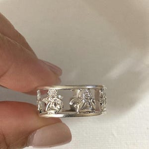 May include: A silver-toned metal ring featuring a repeating cutout design of cherubs or winged figures. The band is constructed with a top and bottom rim, with the figures positioned between the two horizontal bars in a continuous pattern.