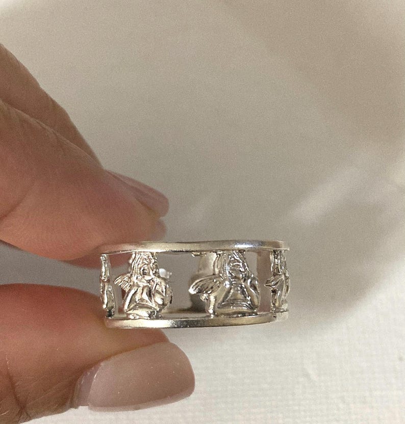 May include: A silver-toned metal ring featuring a repeating cutout design of cherubs or winged figures. The band is constructed with a top and bottom rim, with the figures positioned between the two horizontal bars in a continuous pattern.