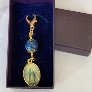 May include: A gold-colored charm with a blue bead and an oval pendant depicting a religious figure. The charm is in a purple box with a dark brown lid. The pendant has a gold border.